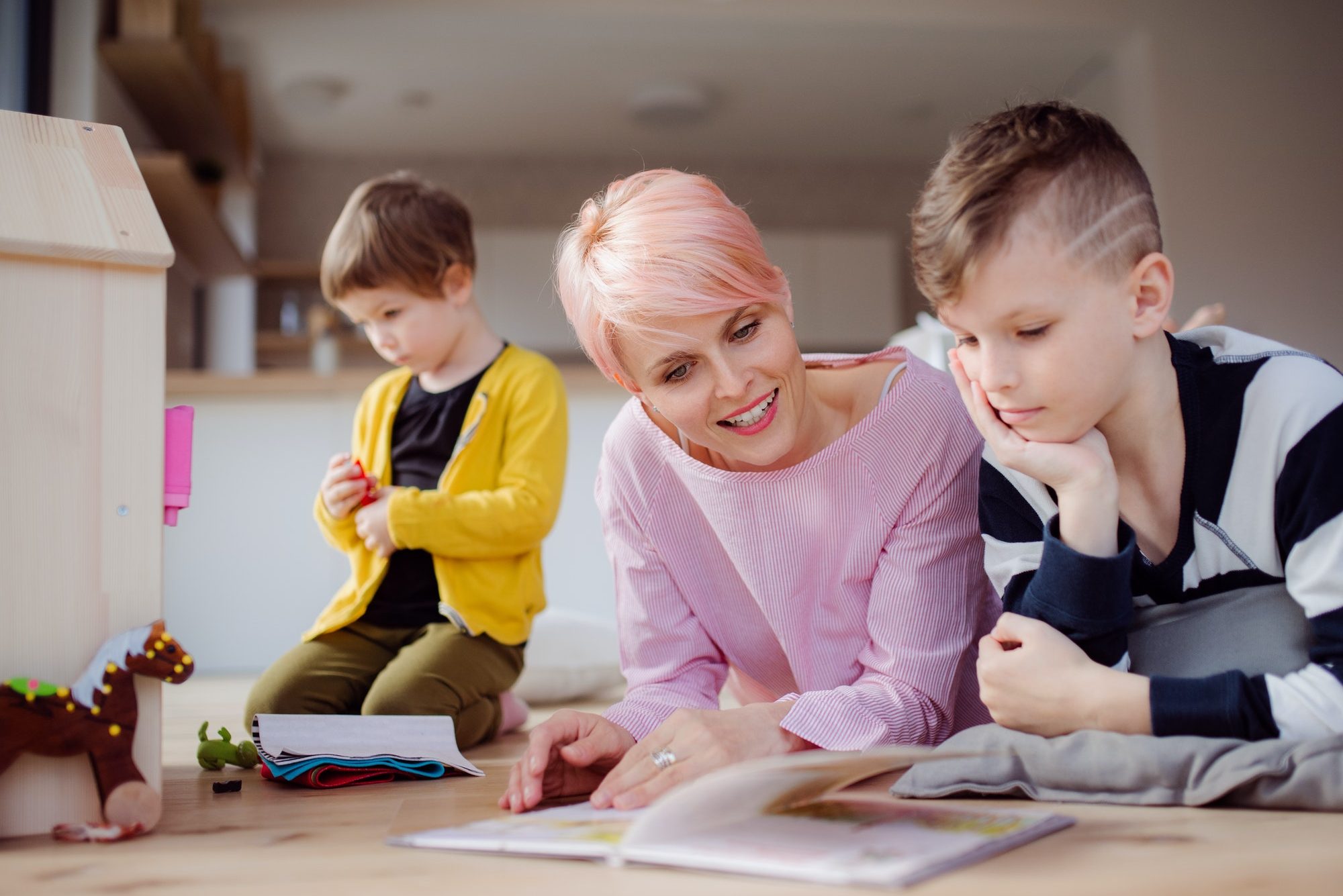 a young woman with two children reading book and playing on the floor e1619662120845