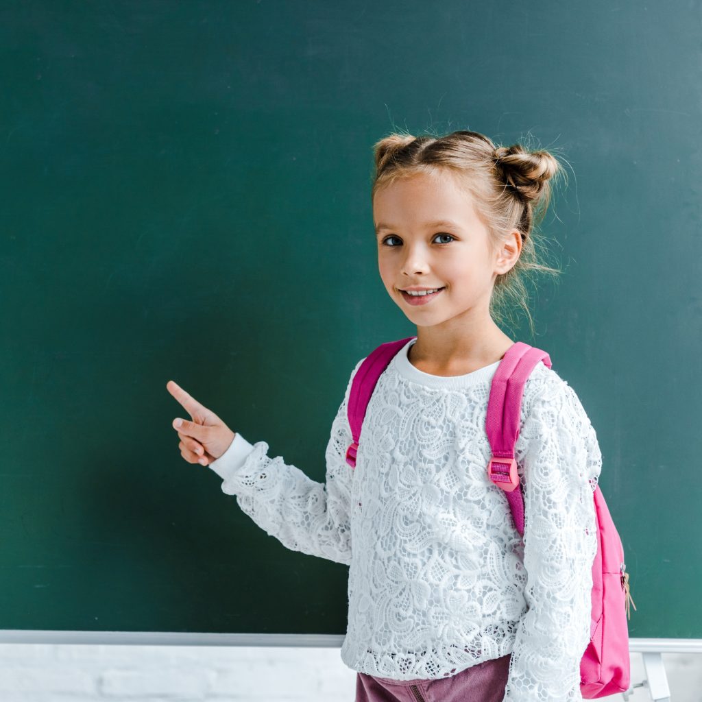happy kid smiling while pointing with finger at green chalkboard